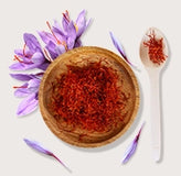 Saffron in a wooden bowl with saffron strands on a spoon, surrounded by saffron flowers.